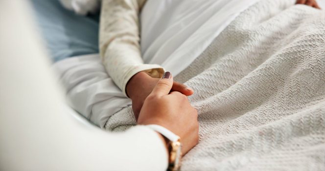 Patient holds hands in hospital bed. Stock image credit / Shutterstock