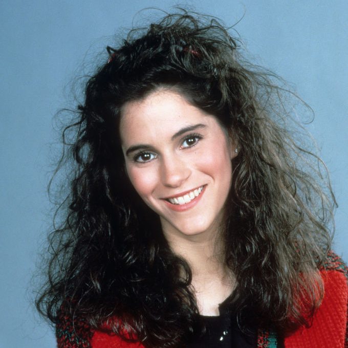 Jami Gertz, w falowanych włosach i czerwono czarno turkusowym swetrze na niebieskim tle