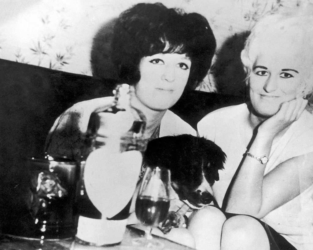 uśmiechnięta Myra Hindley siedzi obok ciemnowłosej kobiety i psa, a na stole stoi alkohol 